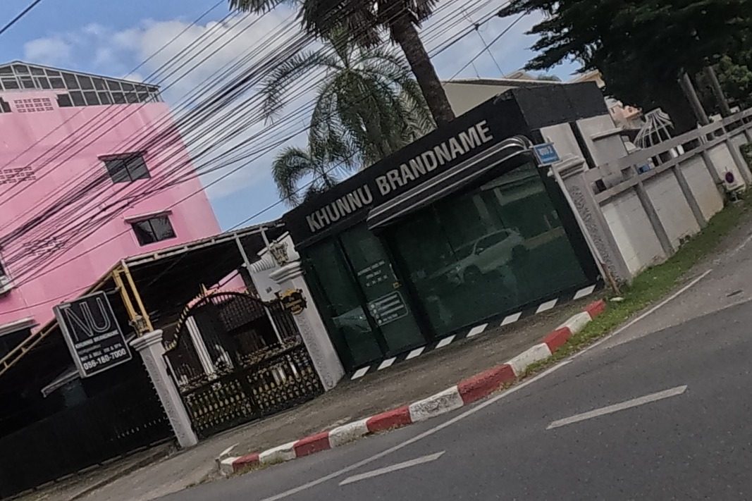 A bright pink building with an NU fashion sign sits beside a branded-goods consignment shop called Khunnu Brandname with a phone number and a car parked inside its glass-fronted showroom.