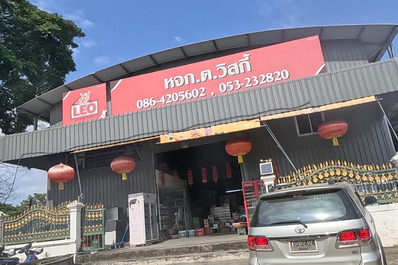 A large beer-wholesale depot, 'หจก.ต.วิสกี้', with LEO and Chang beer branding on its fascia, red Chinese lanterns strung across the entrance, an ornate gilded gate, and delivery pickups marked with the same company name parked outside.