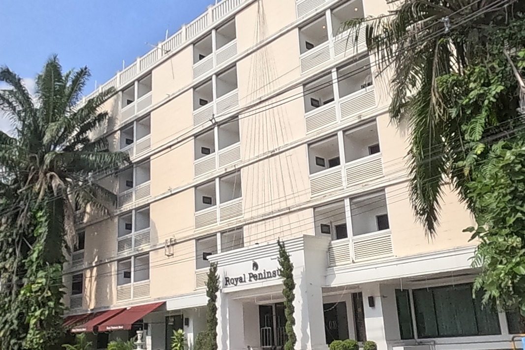 The 'Royal Peninsula' hotel: a six-storey cream building with balconies, palm trees at its entrance, and a small white spirit house with a gilded figure on a pedestal near the driveway; a red-roofed shed and graffiti-marked wall stand across the road.