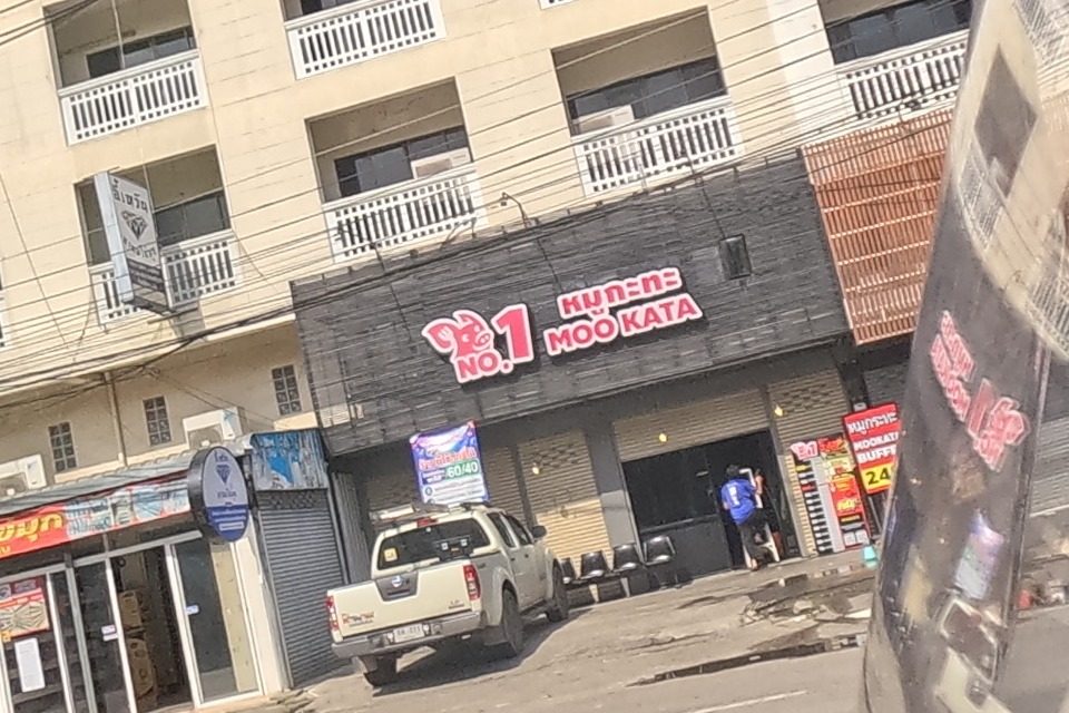 Directly opposite, a 'No.1 Moo Kata' hot-pot restaurant sign with a pig-logo brand mark tops a row of shophouses, next to a shuttered bubble-tea shop and a pickup truck.