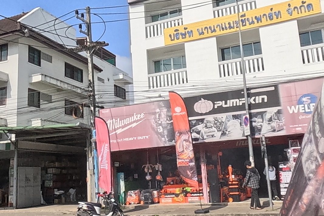 A power-tools shop selling Milwaukee and Pumpkin brands sits next to a welding-equipment shop and a 'Nana Machine Part' company sign, with a Puma store and a 7-Eleven further along the block.