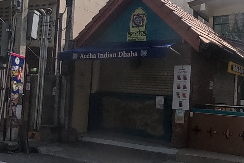 A teal-roofed kiosk signed 'Accha Indian Dhaba' with a colourful 'SPECIAL COMBO' menu board out front; beside it an ornate teal-and-gold gateway leads to a restaurant courtyard with small garlanded shrines and a 'YOGA' banner visible past it.