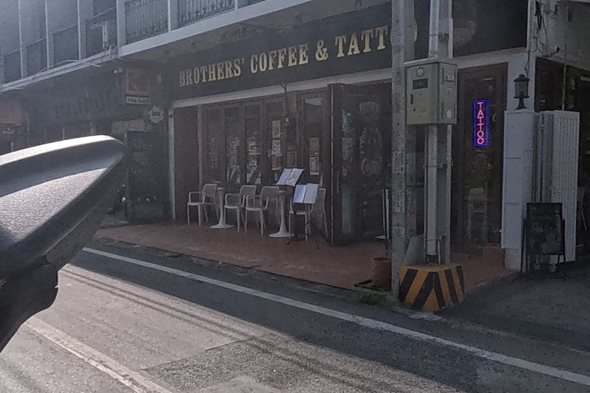A corner cafe and tattoo shop with outdoor seating sits beside a driveway with a small white spirit house on its wall.