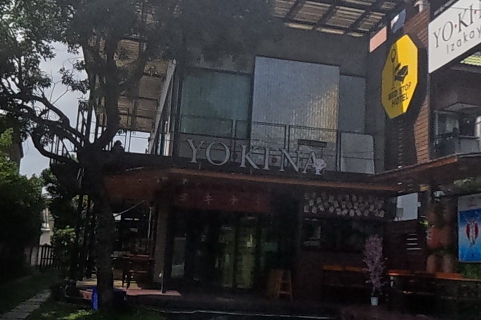A yellow hexagonal Bed Stop Hotel sign and a YO-KI-NA Izakaya signboard mark a wood-trimmed building, beside a small dessert stall with a chest freezer.