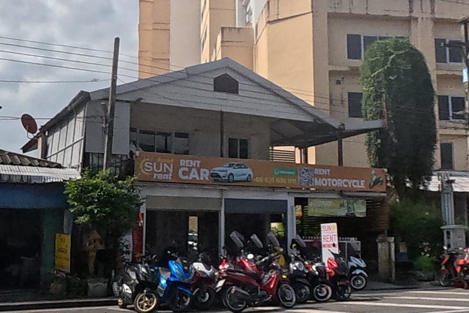 A car-and-motorcycle rental shop with an orange 'SUN rent' sign has scooters lined up outside, beside a tall apartment tower.
