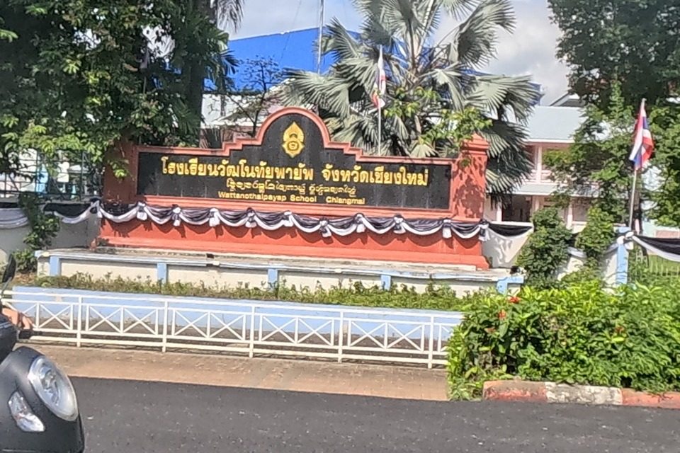 Two women on a scooter pass a black-and-gold monument sign reading Wattanothaipayap School Chiangmai, set among palm trees behind a white picket fence.