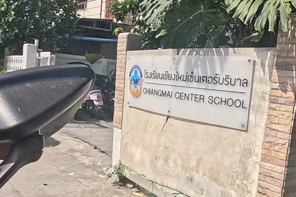 A brick-pillared sign reads “โรงเรียนเชียงใหม่เซ็นเตอร์บริบาล CHIANGMAI CENTER SCHOOL” beside a leafy gate; the school's blue signboard and building continue behind a black car parked in the driveway.
