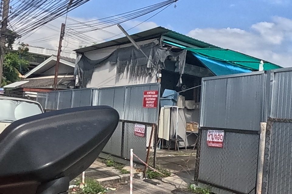 A red sign on a corrugated metal gate reads “VINTAGE SECOND HAND CLOTHING MARKET BICYCLE”, with old furniture and bicycles glimpsed through the gap; a green tarpaulin awning stands beside it.