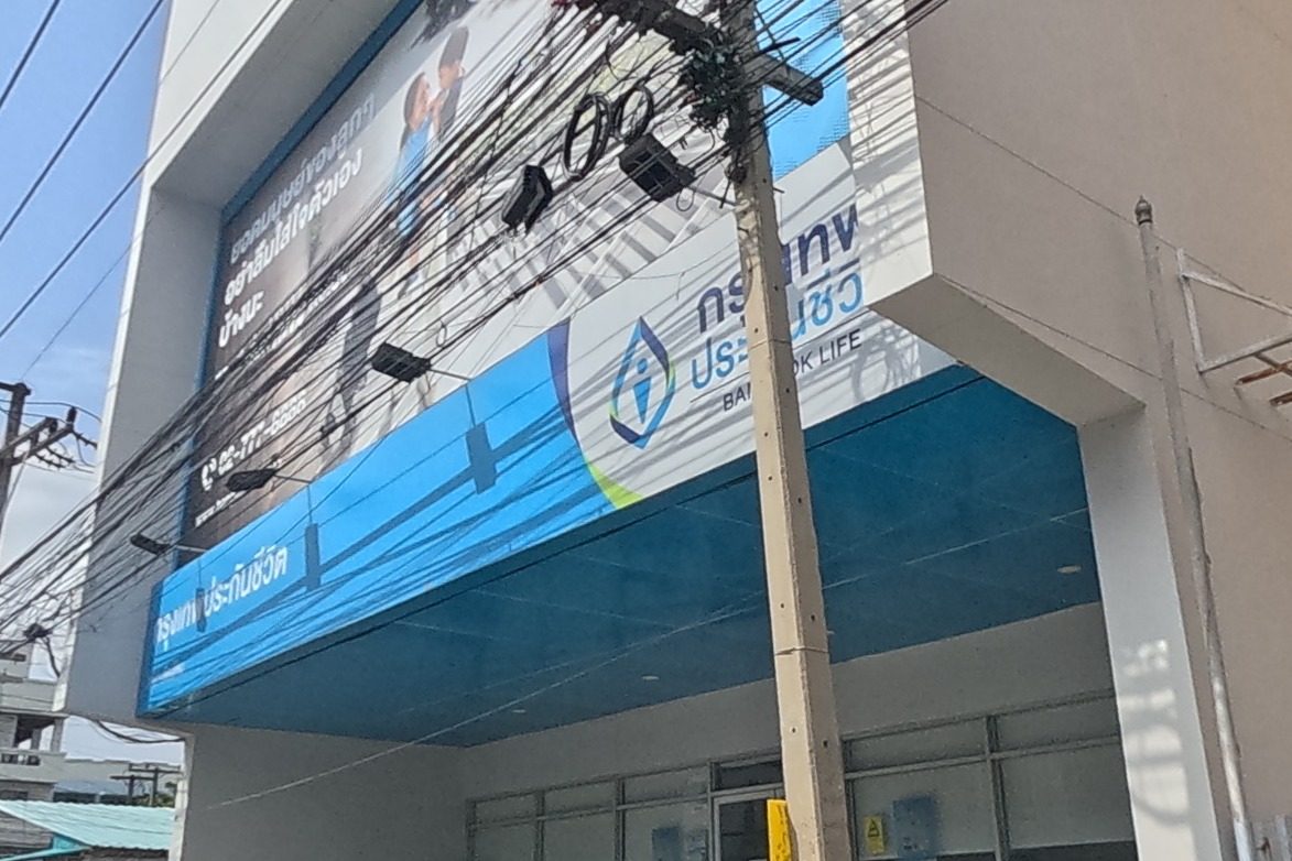 A Bangkok Life Assurance branch office with a blue glass frontage and a matching banner sits beside a service lane leading to parking; motorbikes are parked near the entrance and an orange traffic cone marks the corner.