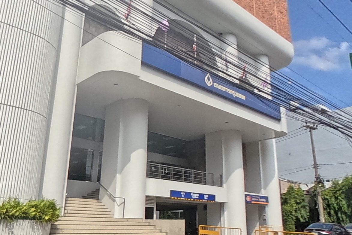 A modern Bangkok Bank branch with tall white columns and blue signage stands on the corner, with an Express Banking self-service kiosk sign at street level and traffic ahead.
