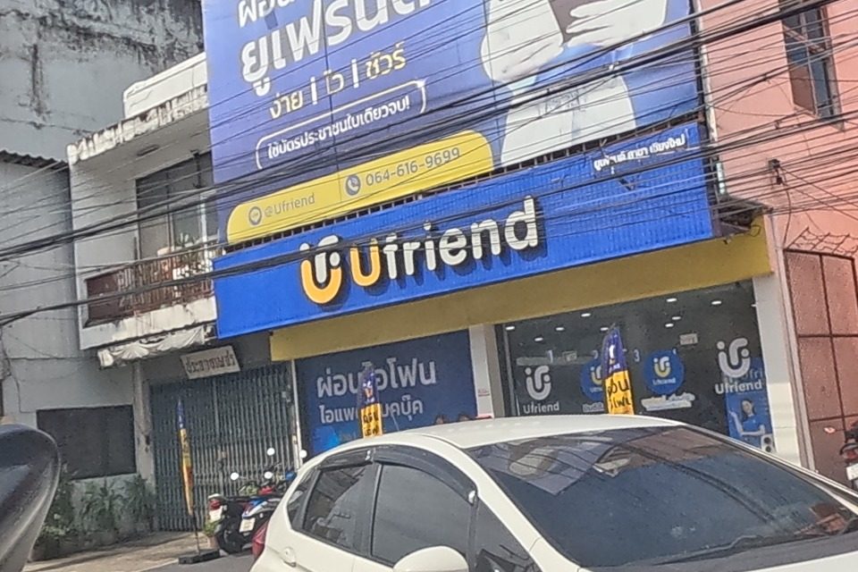 A mobile-phone shop called Ufriend advertises iPhone instalment plans with a LINE ID and phone number, beside a hardware shop selling nuts, screws and tools under an orange sign; a white Honda Jazz drives past.