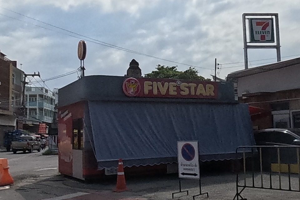 A Five Star long-distance bus company ticket kiosk with a red awning stands beside a 7-Eleven sign and a round pavilion-roofed songthaew stop; traffic cones and a barrier mark off part of the road.