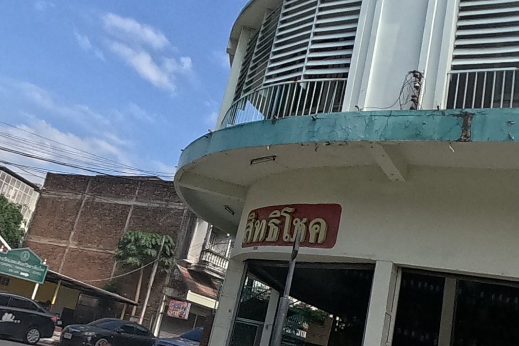 A curved corner building with a red 'Sittichoke' sign houses a shop stacked with sacks and factory-parts equipment, opposite parked sedans; a small roadside booth and a guardhouse stand across the street.
