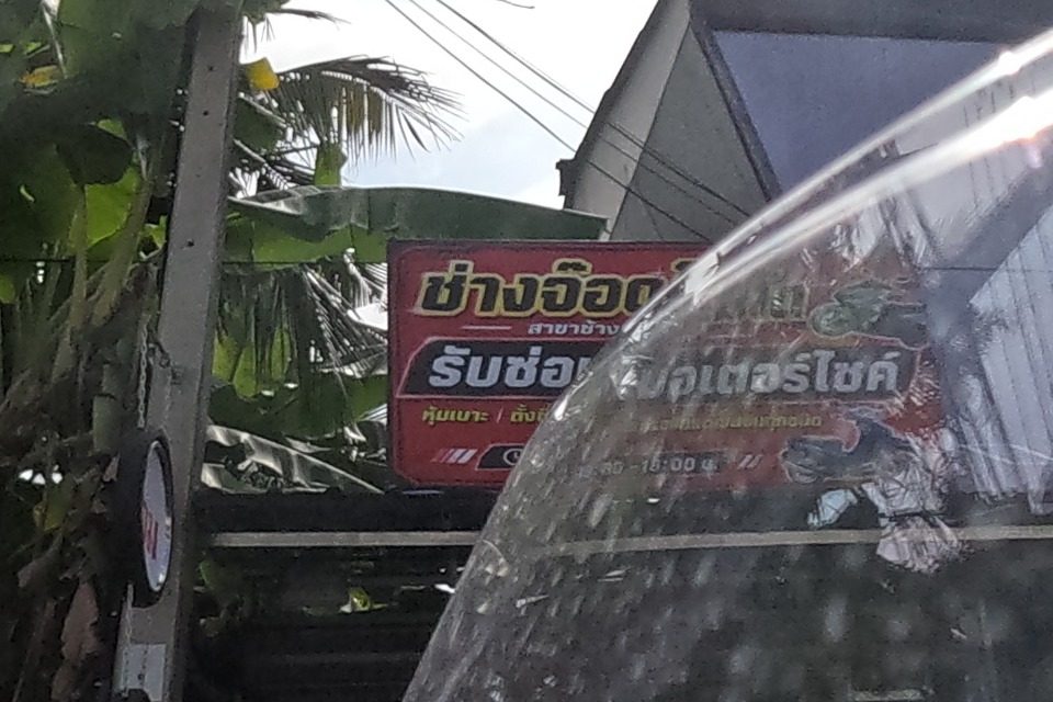 A red sign for a motorcycle-repair shop reading 'ช่างอ๊อด...รับซ่อม...มอเตอร์ไซค์' stands among banana trees, with stacks of old tyres at its base.