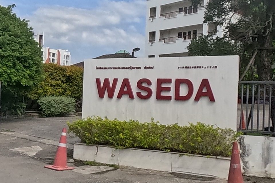 A large white WASEDA sign with red lettering stands behind hedges at the school's street-corner entrance, next to a red anime-style banner.