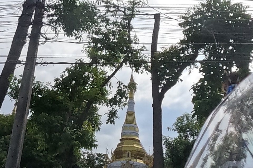 A gilded guardian statue tops the temple's white boundary wall, with the melon-ribbed golden chedi of Wat Ku Tao rising above the trees behind; a tuk-tuk taxi and scooters are parked at the curb.