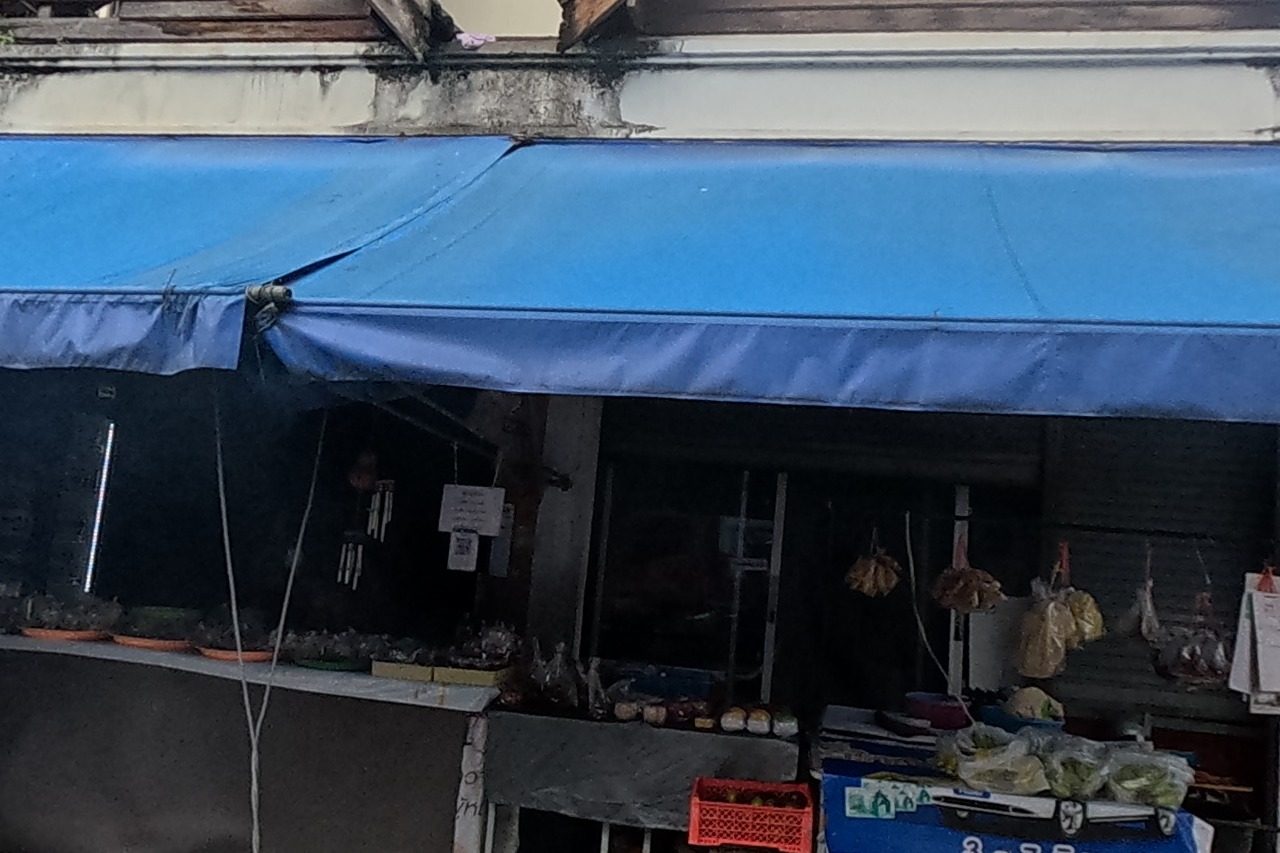 A fresh-produce grocery stall with a blue awning displays vegetables and hanging dried goods under a sign giving dates '3-7 มิ.ย. 65' and phone 053-282-222, beneath an old wooden gable.