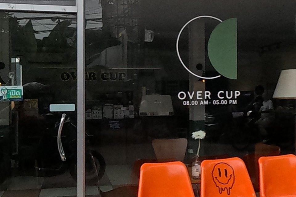 A dark-grey corner cafe called 'OVER CUP' (open 08.00 AM-05.00 PM, unit 29/1) has orange bench seating with a smiley-face decal outside its glass frontage.