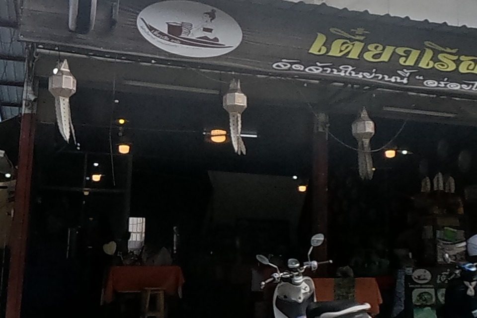 A white 'HELLO' milk-tea kiosk stands beside a boat-noodle restaurant hung with boat-shaped lanterns and a round sign showing a noodle-boat vendor, scooters parked at its entrance.