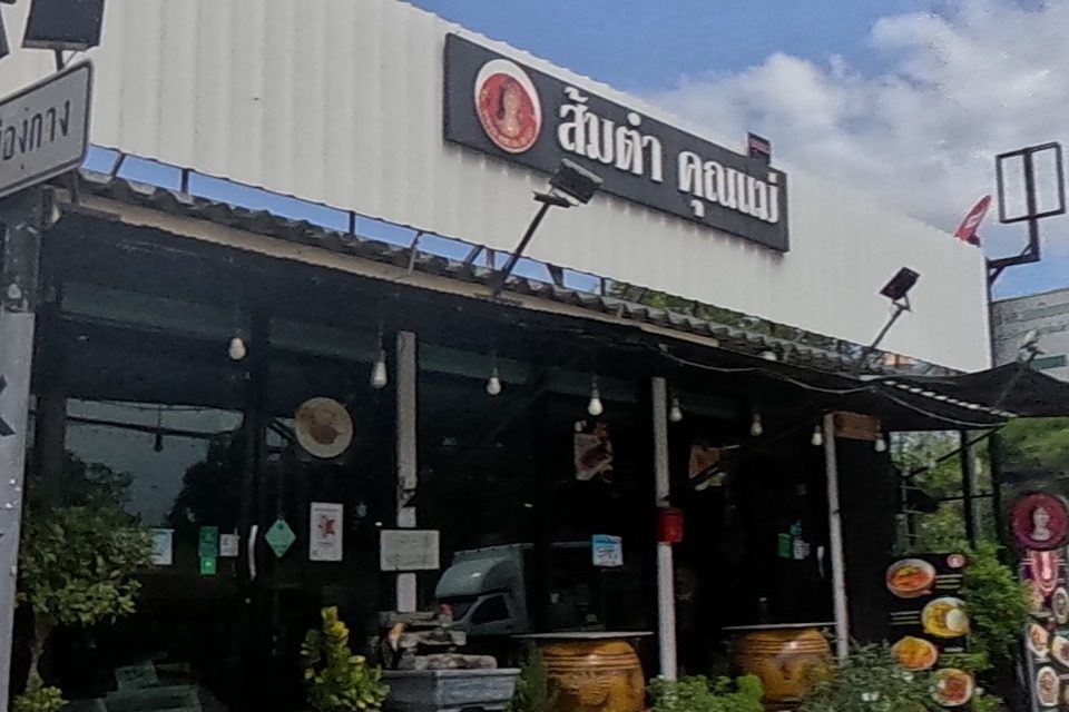 A som-tam restaurant 'ส้มตำ คุณแม่' with a round red logo sign and potted plants at its entrance sits at a green-light intersection, a water tower visible beyond.