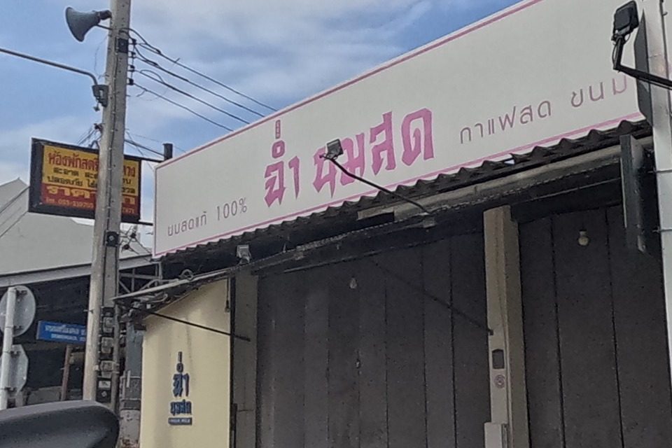 A fresh-milk shop 'ฉ่ำ นมสด' (100% fresh milk, iced coffee, snacks) sits beside 'VINTAGE | UNIQUE STORE' (Harvest Moon), its window lit with an 'OPEN' neon sign and vintage bric-a-brac on display.