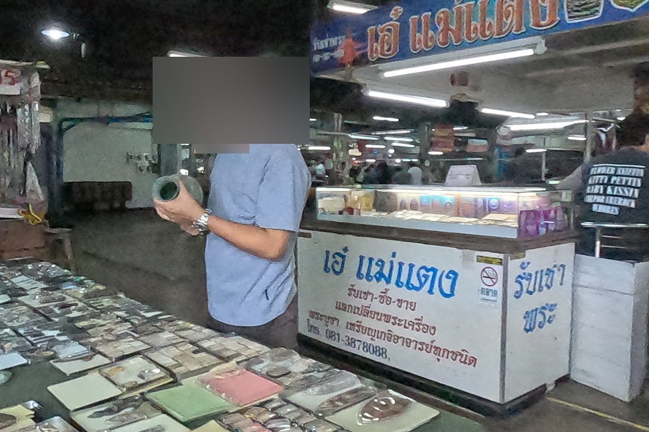 Dealers at a green table laid with open tins of clay and metal amulets, beside the เอ๋ แม่แตง counter with its full sign and phone.