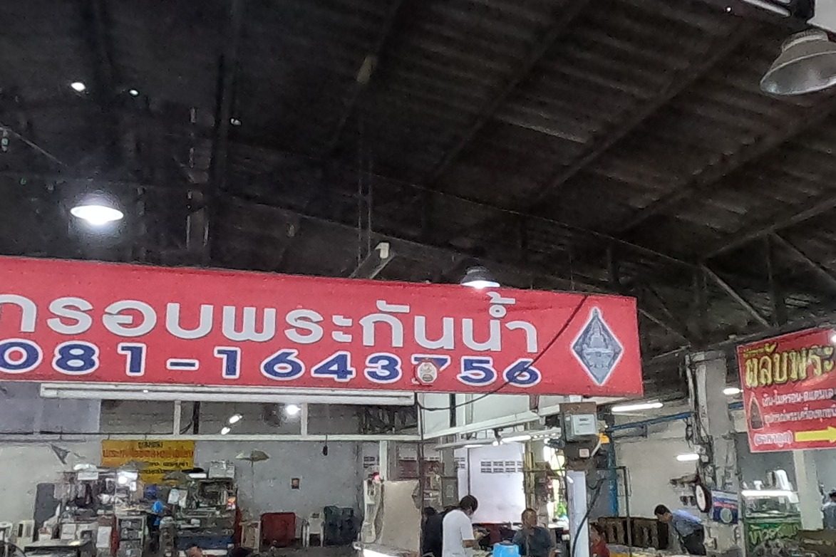 Under the big red หนุ่ม กรอบพระกันน้ำ sign, dealers examine trays of amulets; the hall busy beyond.