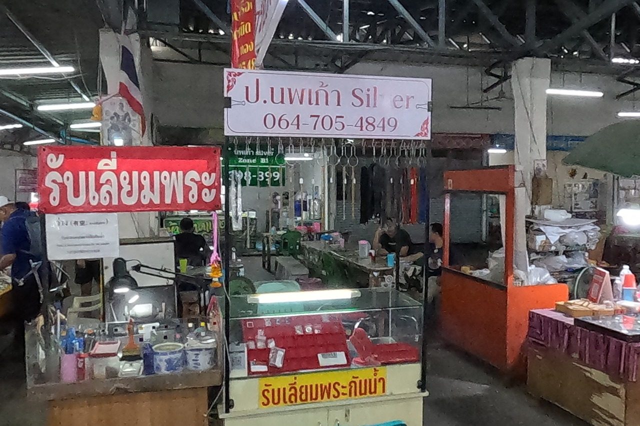 The ป.นพเก้า Silver casing stall with its white sign and phone, red 'รับเลี่ยมพระ' banners and a Thai flag; trays of amulets on a red table in front.