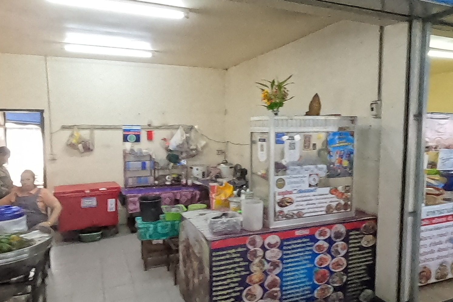 The อิ่มอร่อย โภชนา stall with its menu board and a small shrine shelf; a woman carries a tray of food past the next stall, ร้านแกงเมือง.