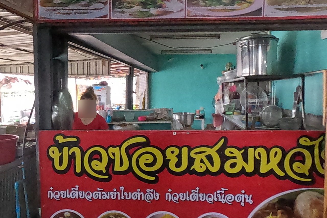 NaN's head in front of the khao soi shop: a woman works at the stove behind the red sign and a numbered photo menu.