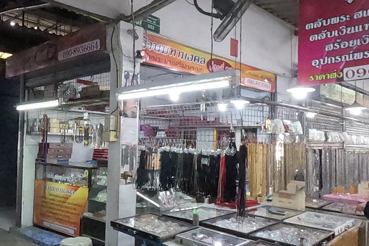 A passage opens between ร้านพระเครื่องตลาดคำเที่ยง and ร้านมหามงคล; glass cases of cases and racks of chains line the shop.