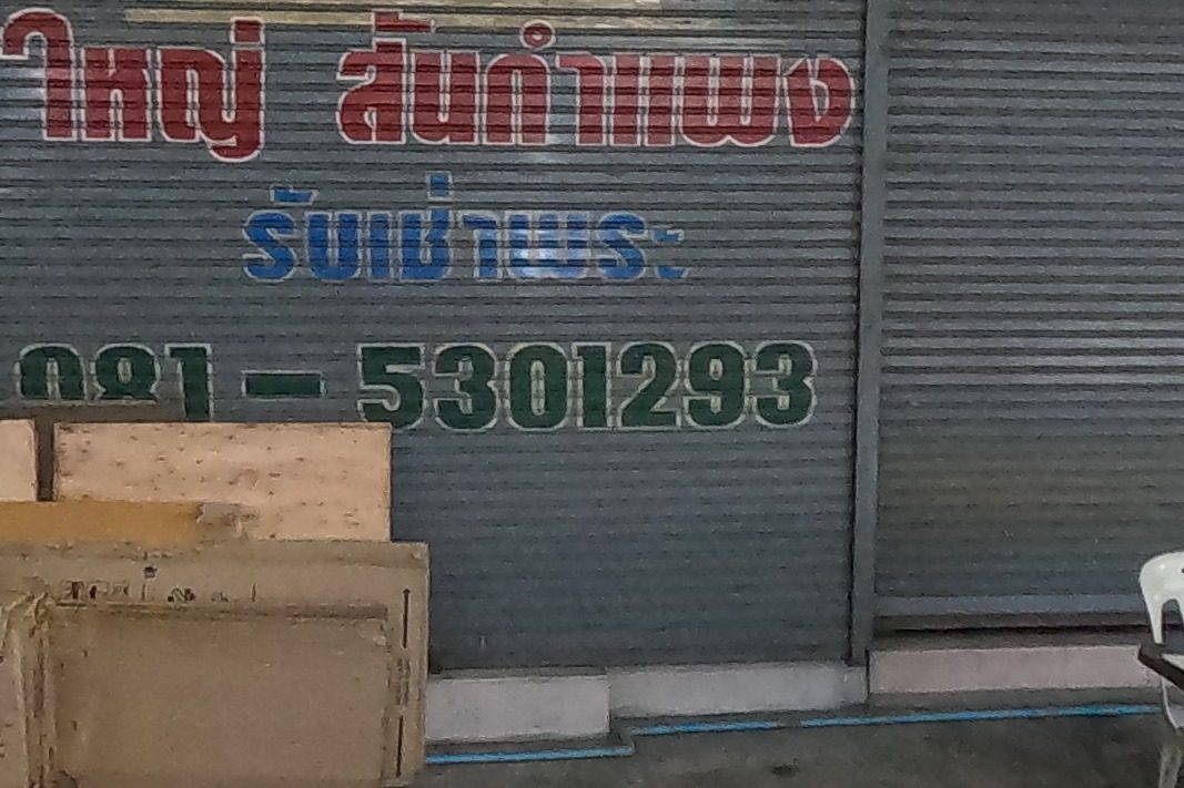 ใหญ่ สันกำแพง's boards and painted shutter beside the black-fascia shop พระเครื่อง ปุ๊ก with its gold lettering; a folding table of old amulets in front, NaN at the lens.