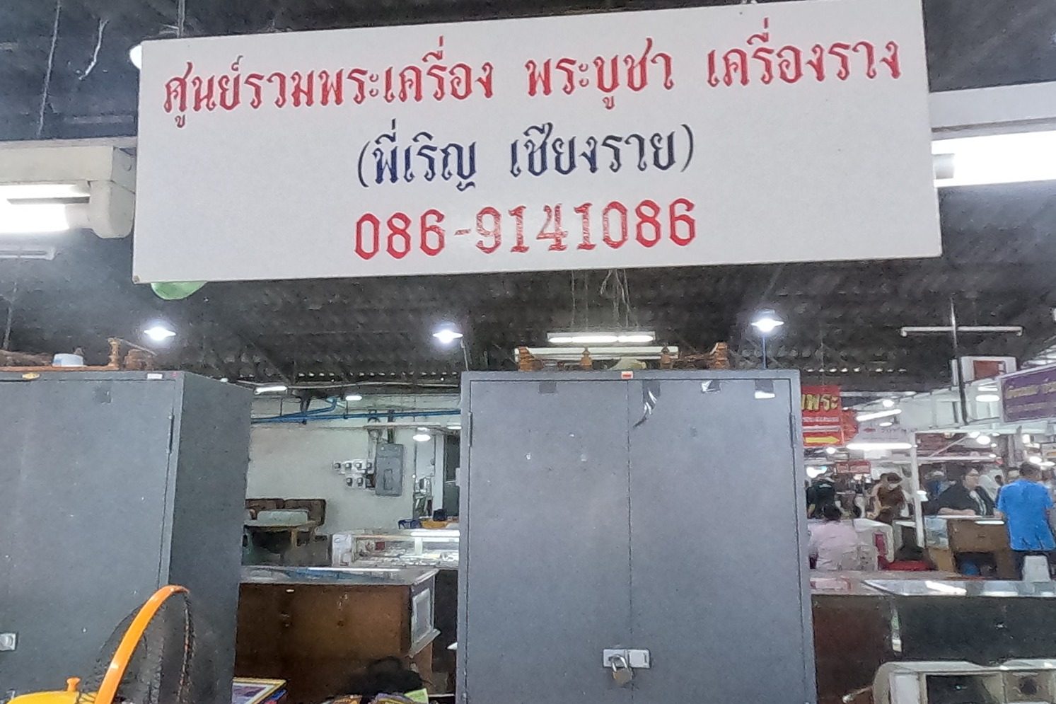 The white sign of ศูนย์รวมพระเครื่อง พระบูชา เครื่องราง (พี่เริญ เชียงราย) with its phone number hangs over two steel cabinets and glass counters; a dealer in purple holds a loupe.