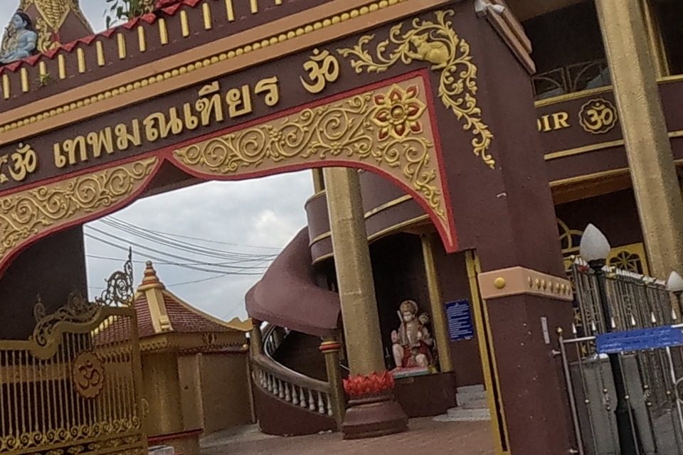 The ornate maroon-and-gold gateway of the Dev Mandir Hindu temple (Thep Montien), with Om symbols, deity statues on top, a gold gate, and a 'please take off your shoes' sign.