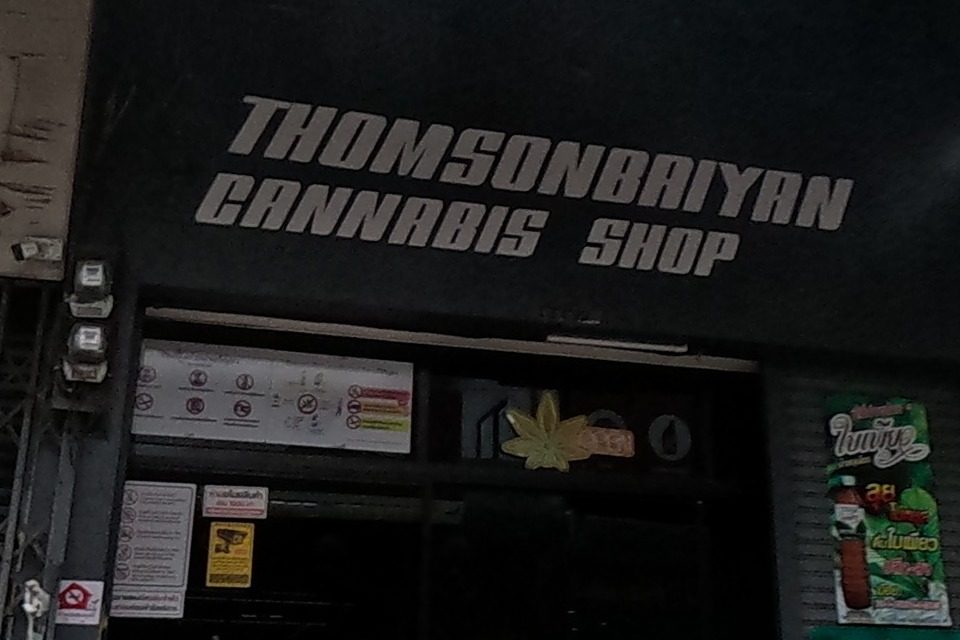A closed shophouse with a faded sign and a 'No Parking' notice, next to Thomsonbaiyan Cannabis Shop, open, with a leaf logo and scooters parked outside.