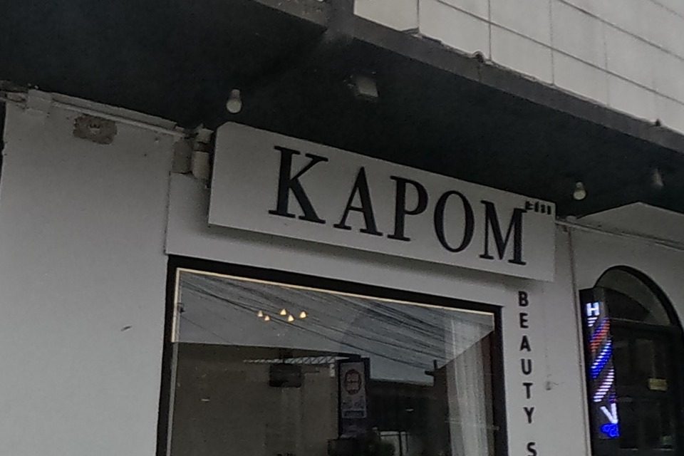 KAPOM Beauty Salon on a street corner, white building with a green painted kerb and a menu signboard by the gate, black cars parked outside.