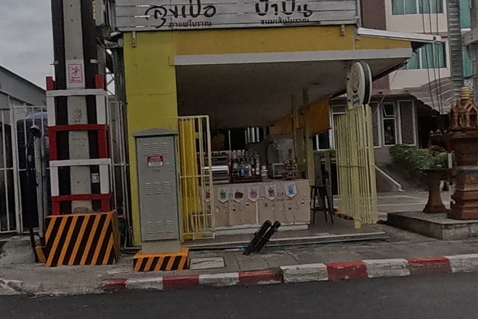 Beside the Power Buy electronics store (with Samsung, LG, Haier, Toshiba and Hisense brand signs), a small yellow corner shop signed 'ลุงเปื้อ ป้าปั้น' sells traditional food and displays several miniature spirit-house models on its step.