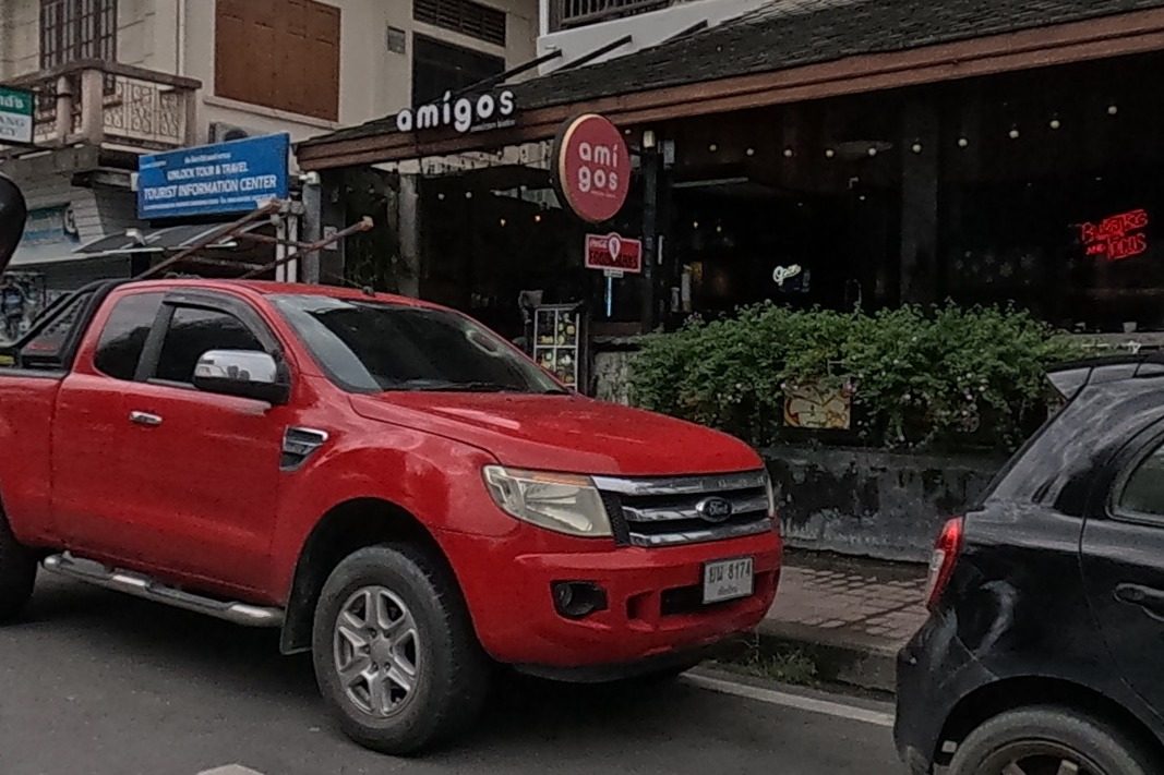 A Mexican restaurant with a neon sign sits under a traditional wooden roof, cars parked outside.