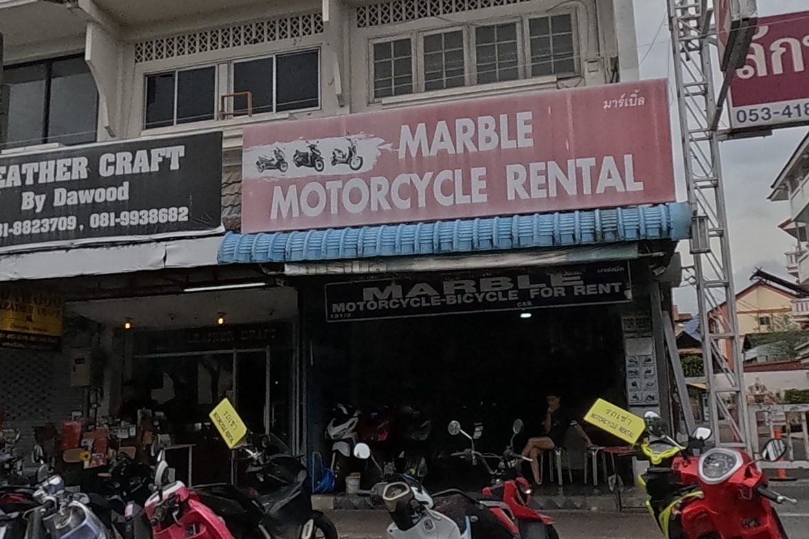 A leather shop, a motorbike rental and a hotel and clinic sign at the corner, scooters parked in front.