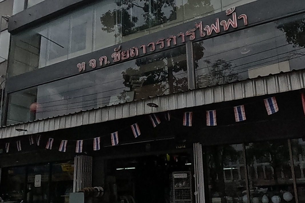 An electrical supplies shop decorated with strings of Thai flags, two pedestrians crossing with an umbrella.