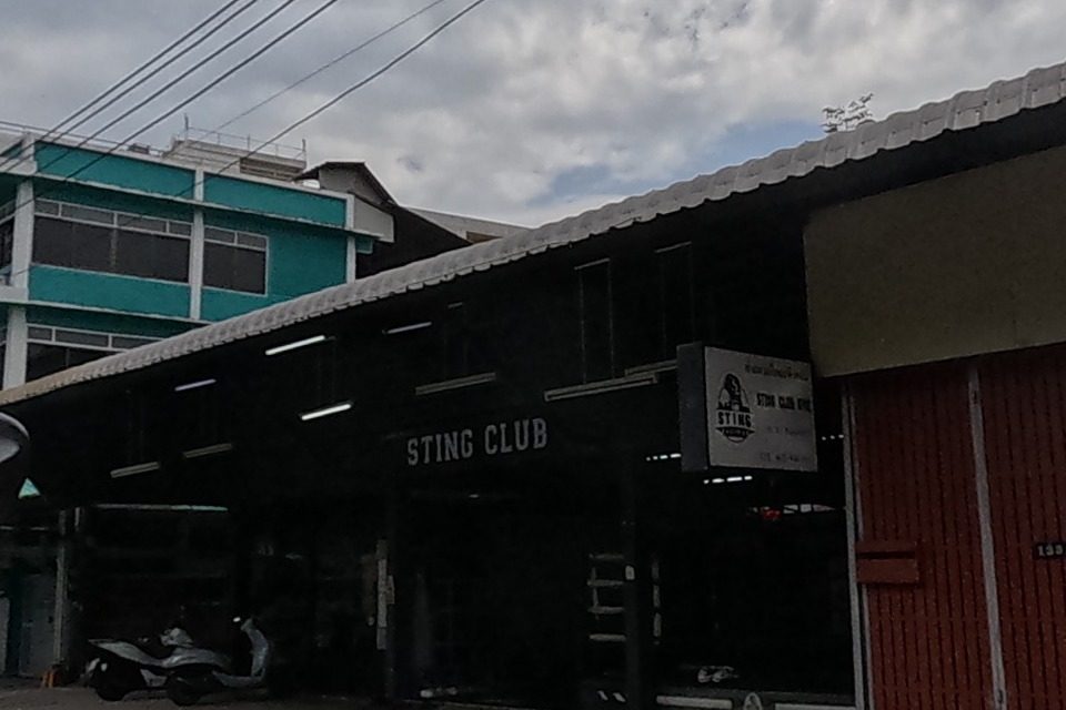 A dark open-sided building with a 'STING CLUB' sign and a shuttered orange roller door sits beside a modern black-and-white townhouse.