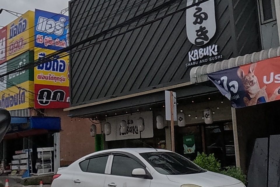 A shabu-and-sushi restaurant sits beside a building with a blue sign advertising a clinic or shop, hung with orange Roojai insurance banners.