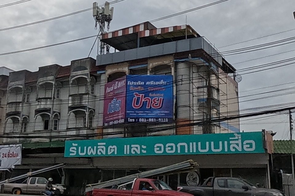 Shophouses across the median: a garment/sign printing shop 'รับผลิต และ ออกแบบเสื้อ' with 'โปรดัค ครีเอชั่น' banners advertising shirts and signage above it.