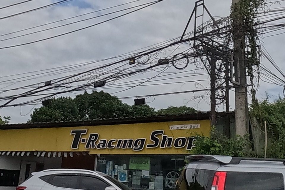 T-Racing Shop, a motor-parts shop with a yellow awning, two SUVs parked outside; a side road leads off to housing-project billboards ('LA VILLETTA' and another) beyond.