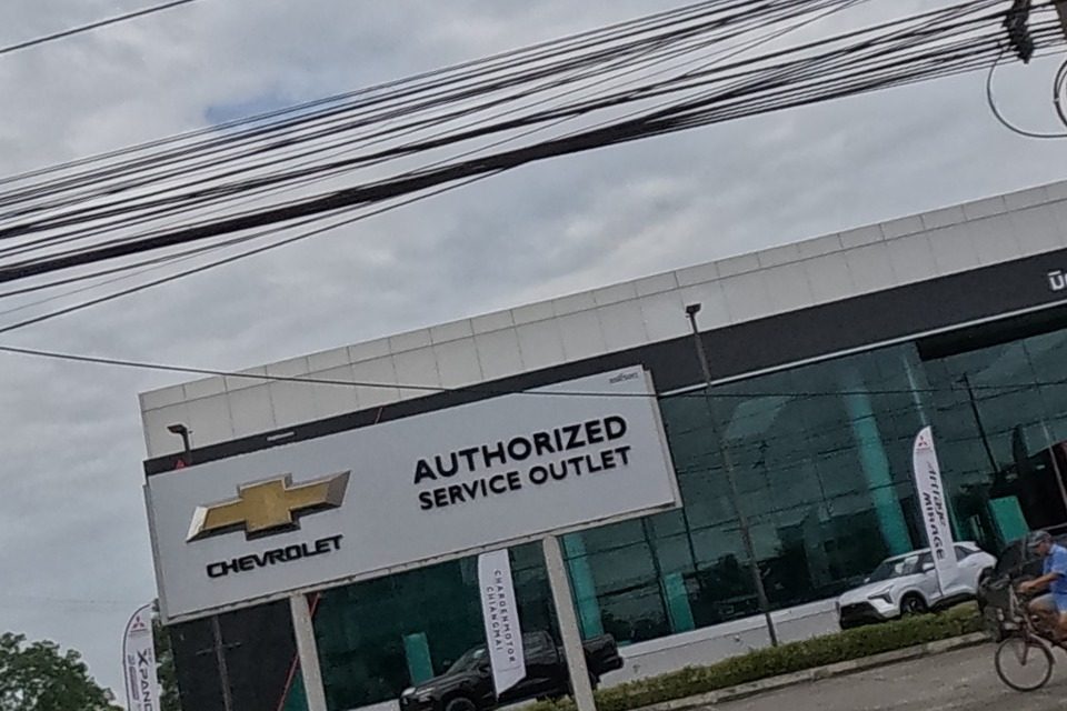 A Mitsubishi Motors pylon sign and, behind it, a Chevrolet dealership with an 'Authorized Service Outlet' sign, a black pickup on display, and a cyclist crossing the forecourt.