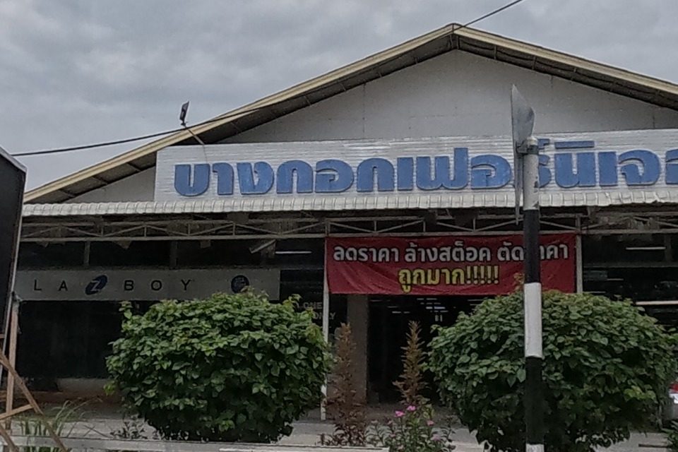 A furniture store 'Bangkok Furniture' (stocking La-Z-Boy) with a red banner advertising a clearance sale, and a yellow price sign for a car-wash/detailing service (50-70% off) next door.