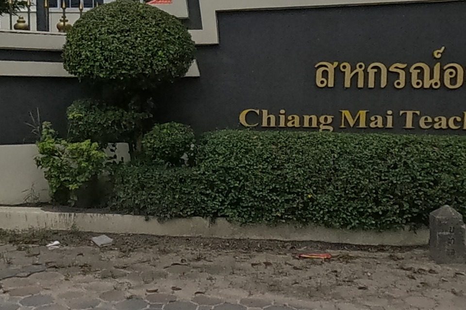 A stone sign at a gated compound reading 'Chiang Mai Teacher's Thrift & Credit Cooperative Ltd.' in Thai and English, with a modern building behind.