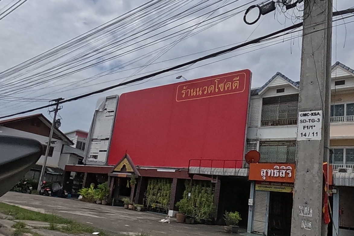 The Chokdi massage shop's red sign, a small shop below it, a gated compound next door with a marigold-print privacy banner and a land-for-sale sign with a phone number, townhouses behind.