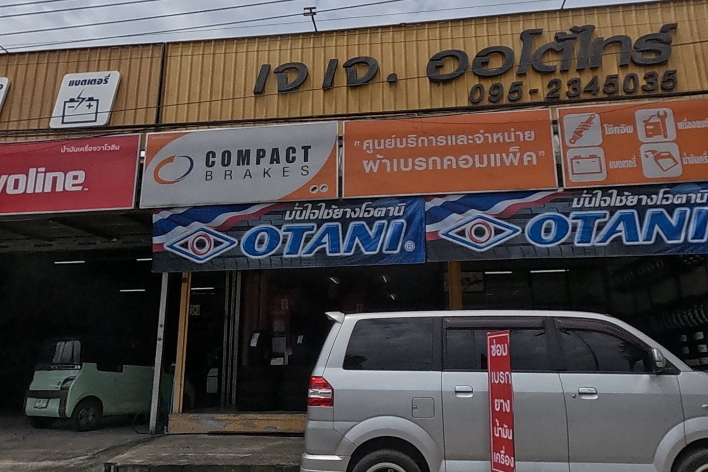A tyre and auto-service shop with Valvoline, Compact Brakes and Otani tyre banners, phone number on its main sign, a silver van parked out front, directional signs to the train station and Sankamphaeng district.