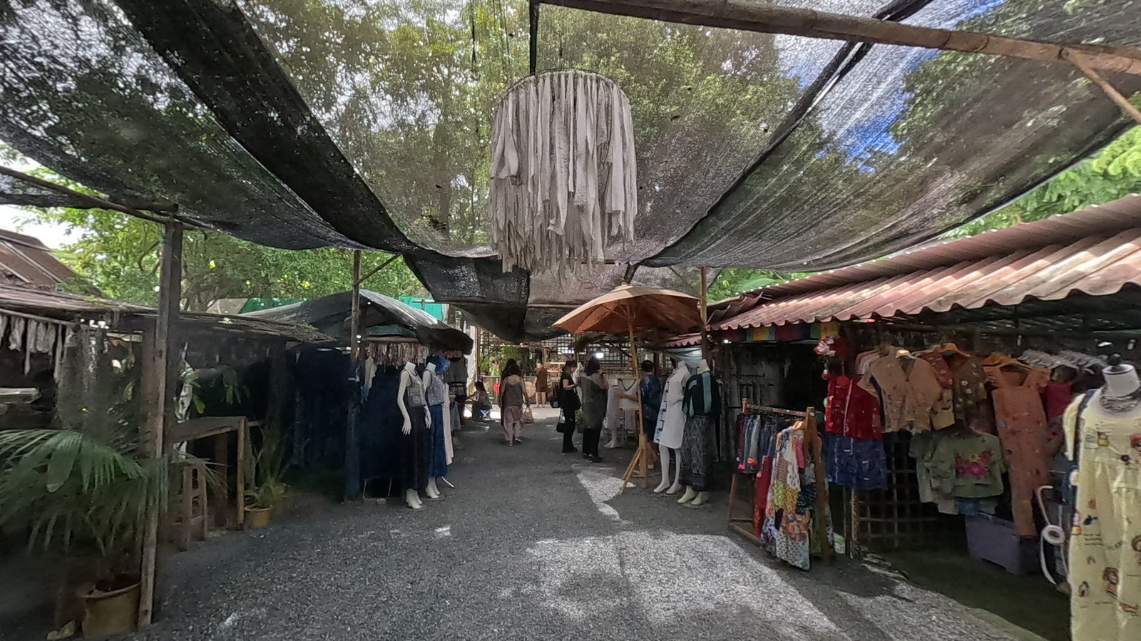 The clothing lane under shade nets and a cloth-strip lantern — Chamcha Market, San Kamphaeng
